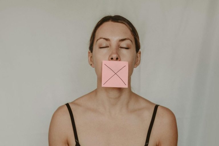 Une femme en buste, qui ferme les yeux et qui a sur la bouche un post it marqué d'une croix